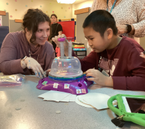 Educator Taylor Knouse-Selk sitting at a desk with a young male student that is using a modified version of the spin art toy.