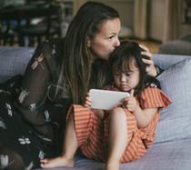 A mother kissing her daughter on the top of their head while sitting on the couch.