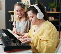 A teenage student wearing a yellow sweater is playing the keyboard next to an educator.