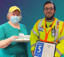 Two people at work. One is a woman holding a cooking tray and the other is a man wearing a safety vest holding a certificate of recognition.